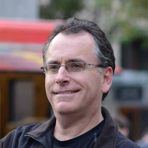 A middle-aged man with short dark hair and glasses smiles outdoors. He is wearing a black jacket and a black shirt, with a blurred background featuring a red structure and greenery.